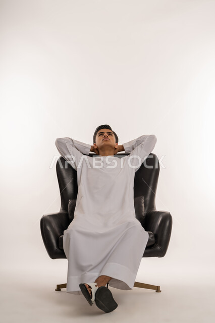 Feeling tired and needing to sleep, looking at something with expressions of thinking and absent-mindedness, the concept of relaxation and enjoying the calm atmosphere, a portrait of a young Arab Gulf Emirati teenager wearing a white kandura sitting on a comfortable chair with his hands behind his head, white background
