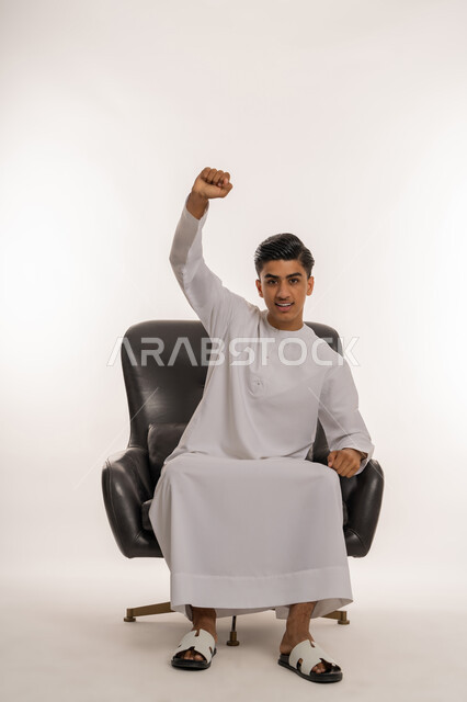 تحقيق الاهداف و إنجاز المهام, الجلوس على كرسي مريح والنظر الى الكاميرا بإيماءات السرور, علامات وتعابيرالفرح والقوة والرضا، بورتريه لشاب مراهق عربي خليجي اماراتي مبتسم يرتدي الكندورة الاماراتية يرفع قبضة يديه بإيماءات النصر والفوز و الكسب, خلفية بيضاء
