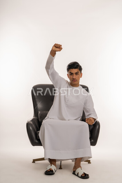 Sitting on a comfortable chair and looking at the camera with serious gestures, achieving goals and accomplishing tasks, expressions of strength and satisfaction, portrait of a young Arab Gulf Emirati teenager wearing an Emirati kandura raising his fists with gestures of victory, winning and gain, white background