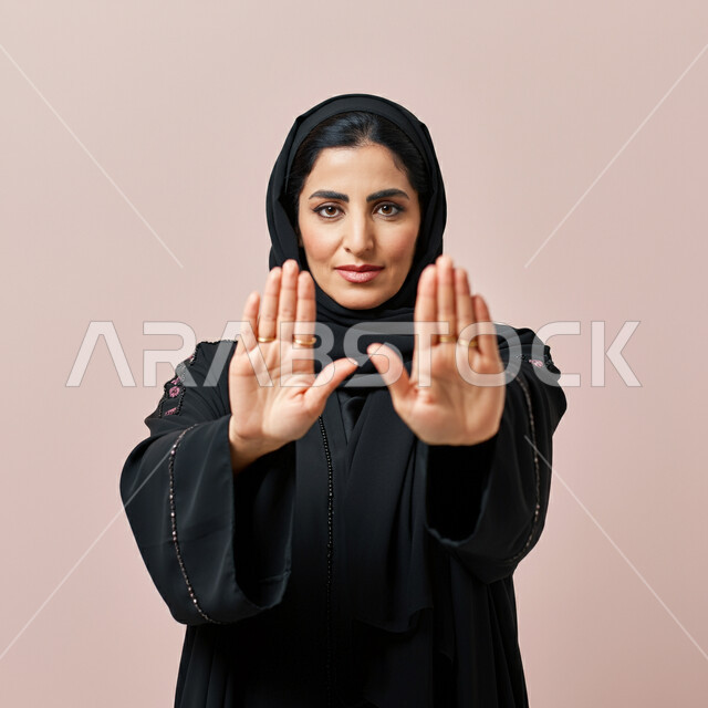 الأمر بالتوقف عن فعل شيء ما، الشعور بالإنزعاج والغضب، تعابير الرفض والإمتناع، بورتريه مقرب لامرأه عربيه خليجيه سعوديه محجبة ترتدي عباءة سوداء ترفع يدها بإيماءات تدل على النهي، خلفية وردية