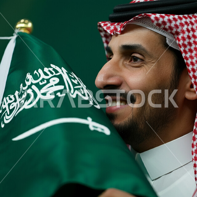 الاحتفال باليوم الوطني لتوحيد السعودية، تعميق الانتماء والولاء للوطن، بورتريه مقرب لشاب عربي خليجي سعودي يرتدي الشماغ والثوب التقليدي يحمل علم السعودية وينظر إلى شيء ما بفخر وتباهي، العيد الوطني السعودي 23 سبتمبر، يوم العلم السعودي 11 مارس، كروما، خلفية خضراء