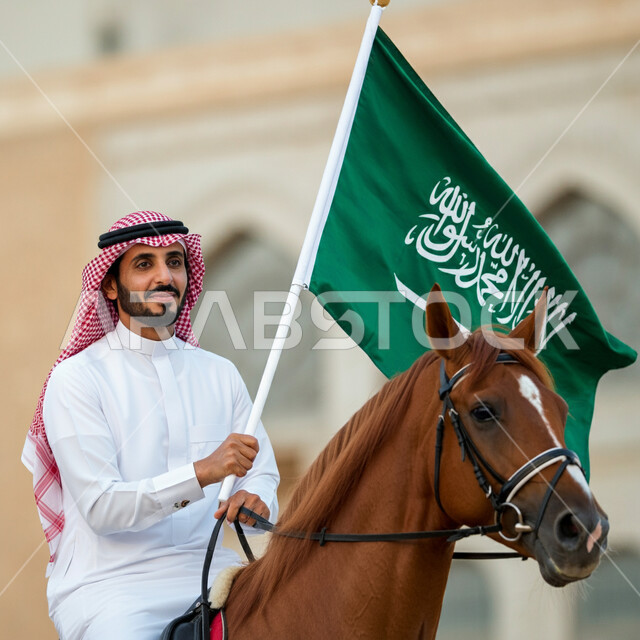 الاحتفال باليوم الوطني لتوحيد السعودية، يوم العلم السعودي 11 مارس، حب الوطن والانتماء إليه، صورة من الجانب لرجل عربي خليجي سعودي يرتدي الثوب التقليدي والشماغ يحمل علم السعودية يركب على الخيل بإيماءات الفخر، العيد الوطني السعودي 23 سبتمبر