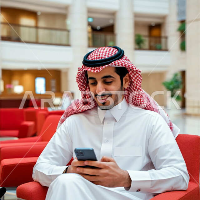 Holding a business meeting online, using modern technical devices, a Saudi Arabian Gulf man wearing a traditional shemagh and thobe sitting in a hotel waiting room holding a mobile phone in his hand, looking at it with gestures of happiness and pleasure, browsing social media sites