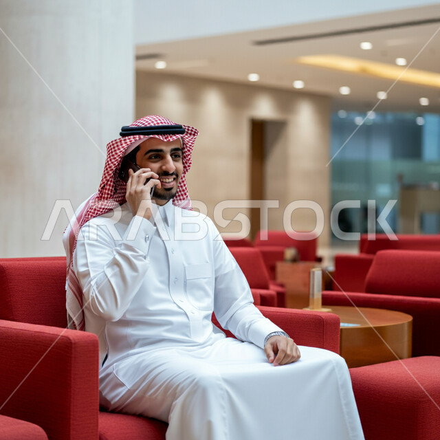 سماع أخبار جيدة من الأهل والأصدقاء، استخدام الاجهزة التقنية الحديثة، رجل عربي خليجي سعودي يرتدي الشماغ والثوب التقليدي يجلس في قاعة انتظار الفندق يقوم بإجراء مكالمة هاتفية عبر الهاتف المحمول، تصفح مواقع التواصل الاجتماعي 
