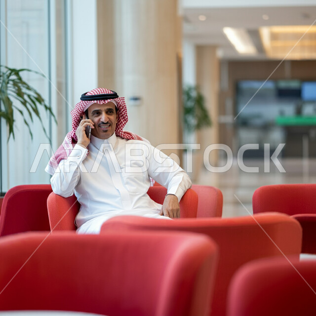 سماع أخبار جيدة من الأهل والأصدقاء، استخدام الاجهزة التقنية الحديثة، رجل عربي خليجي سعودي يرتدي الشماغ والثوب التقليدي يجلس في قاعة انتظار الفندق يقوم بإجراء مكالمة هاتفية عبر الهاتف المحمول، تصفح مواقع التواصل الاجتماعي 