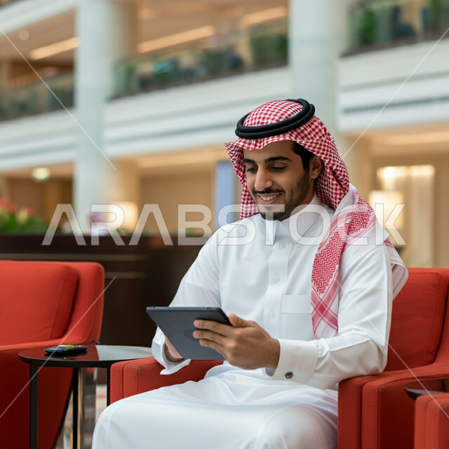 استخدام الاجهزة التقنية الحديثة، رجل عربي خليجي سعودي يرتدي الشماغ والثوب التقليدي يجلس في قاعة انتظار الفندق يمسك بيده الجهاز اللوحي المحمول ينظر إليه بإيماءات السعادة والسرور، عقد إجتماع عمل عبر الإنترنت، تصفح مواقع التواصل الاجتماعي 