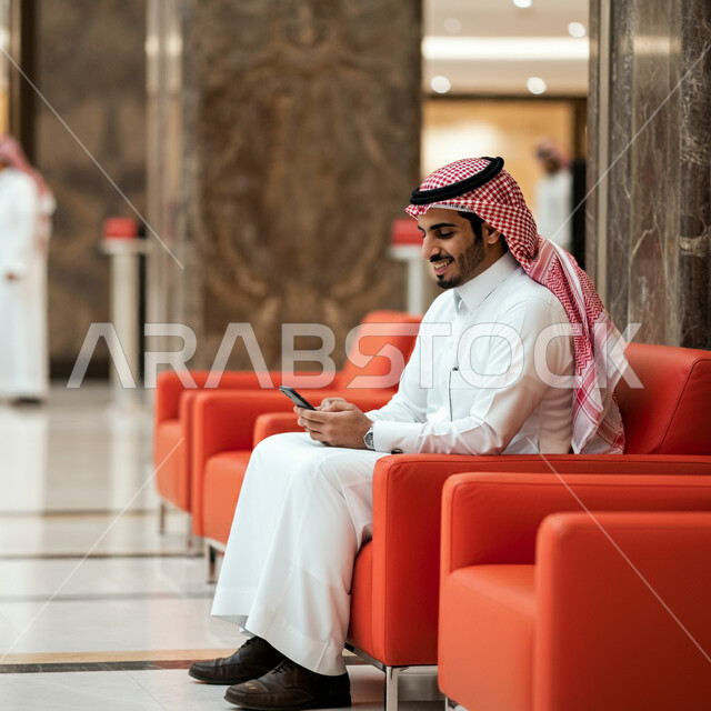 استخدام الاجهزة التقنية الحديثة، صورة من الجانب لرجل عربي خليجي سعودي يرتدي الشماغ والثوب التقليدي يجلس في قاعة انتظار الفندق يمسك الهاتف المحمول، سماع أخبار جيدة من الأهل والأصدقاء، تصفح مواقع التواصل الاجتماعي 
