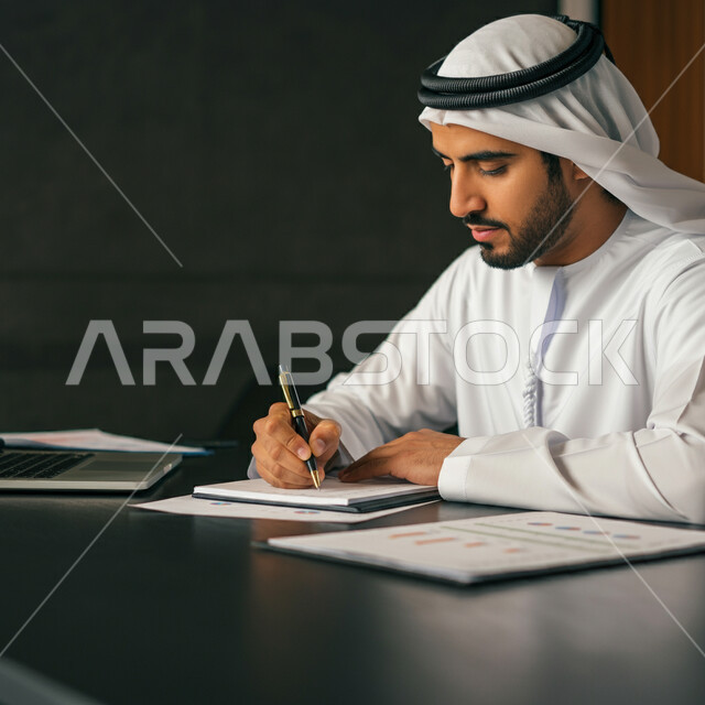 Writing and recording information on paper, close-up image of a young Emirati Gulf Arab man wearing a kandura and ghutra sitting on a chair in his office holding a pen and writing notes, defining daily tasks and goals, the concept of managing and organizing business affairs