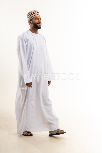 الاهتمام بالأناقة والمظهر الخارجي، مفهوم الرجولة والثقة بالنفس، التقاط الصور الشخصية للوثائق الرسمية، بورتريه لشاب عربي خليجي عماني مبتسم يرتدي الدشداشة و الكمه بإيماءات السرور والسعادة، الجسم بالطول الكامل، خلفية بيضاء