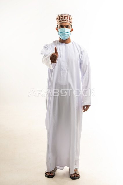 الاهتمام بالاناقة والمظهر الخارجي، اتباع الإجراءات الاحترازية لمنع انتشار العدوى، بورتريه لشاب عربي خليجي عماني يرتدي الدشداشة والكمه يضع الكمامة يرفع اصبعه الابهام بايماءات الرضا والموافقة، اتخاذ التدابير اللازمة للوقاية من فيروس كورونا، الجسم بالطول الكامل، خلفية بيضاء