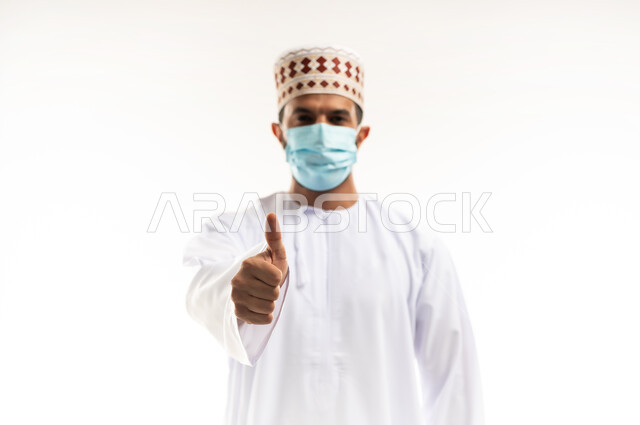 اتباع الإجراءات الاحترازية لمنع انتشار العدوى، بورتريه مقرب لشاب عربي خليجي عماني يرتدي الدشداشة والكمه يضع الكمامة الطبية يرفع اصبع الابهام بايماءات الرضا والاعجاب، اتخاذ التدابير اللازمة للوقاية من فيروس كورونا،  خلفية بيضاء