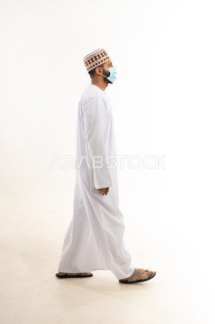 Caring about elegance and appearance, following precautionary measures to prevent the spread of infection, portrait of a young Gulf Arab Omani man wearing a dishdasha and a cuff, wearing a medical mask to avoid infection, taking the necessary measures to prevent the Corona virus, full-length body, white background