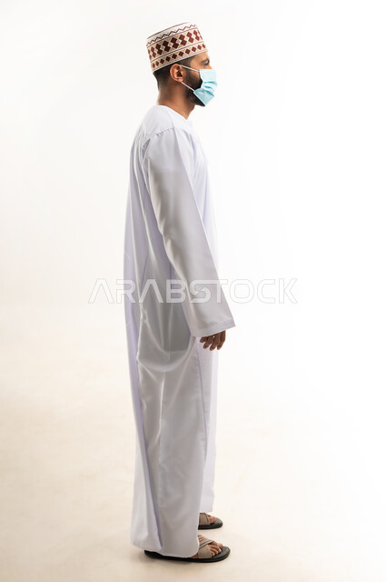 الاهتمام بالاناقة والمظهر الخارجي، اتباع الإجراءات الاحترازية لمنع انتشار العدوى، بورتريه من الجانب لشاب عربي خليجي عماني يرتدي الدشداشة والكمه يضع الكمامة الطبية لتجنب الاصابة بالامراض، اتخاذ التدابير اللازمة للوقاية من فيروس كورونا، الجسم بالطول الكامل، خلفية بيضاء