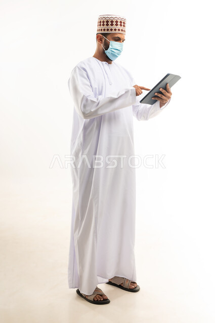 احترام قوانين وقواعد وزارة الصحة، استخدام التقنيات الحديثة المتطورة، بورتريه لشاب عربي خليجي عماني يرتدي الدشداشة والكمه يضع القناع الطبي للوقاية من فايروس كورونا كوفيد-19يحمل جهاز التابليت، الجسم بالطول الكامل،خلفية بيضاء
