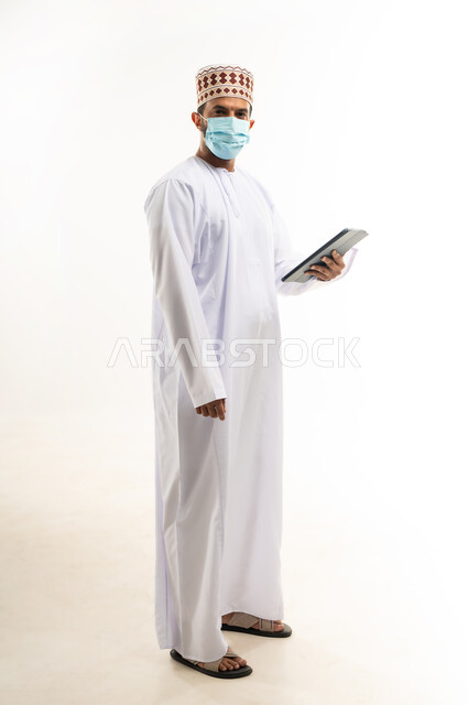 احترام قوانين وقواعد وزارة الصحة، استخدام التقنيات الحديثة المتطورة، بورتريه لشاب عربي خليجي عماني يرتدي الدشداشة والكمه يضع القناع الطبي للوقاية من فايروس كورونا كوفيد-19يحمل جهاز التابليت، الجسم بالطول الكامل،خلفية بيضاء