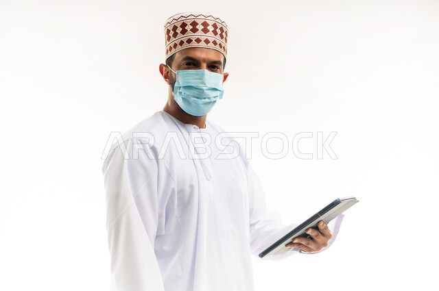 النظر الى الكاميرا بايماءات الجدية، احترام قوانين وقواعد وزارة الصحة، استخدام التقنيات الحديثة المتطورة، بورتريه مقرب لشاب عربي خليجي عماني يرتدي الدشداشة والكمه يضع القناع الطبي للوقاية من فايروس كورونا كوفيد-19يحمل جهاز التابليت، خلفية بيضاء