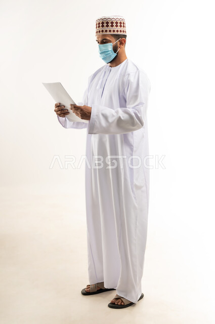الحفاظ على السلامة من كوفيد 19، لوحات توعوية لتجنب إنتشار العدوى، اتباع الاجراءات الاحترازية للوقاية من فايروس كورونا، بورتريه لرجل عربي خليجي عماني يرتدي الدشداشة والكمة يضع الكمامة يمسك أوراق بيضاء فارغة, الجسم بالطول الكامل، خلفية بيضاء
