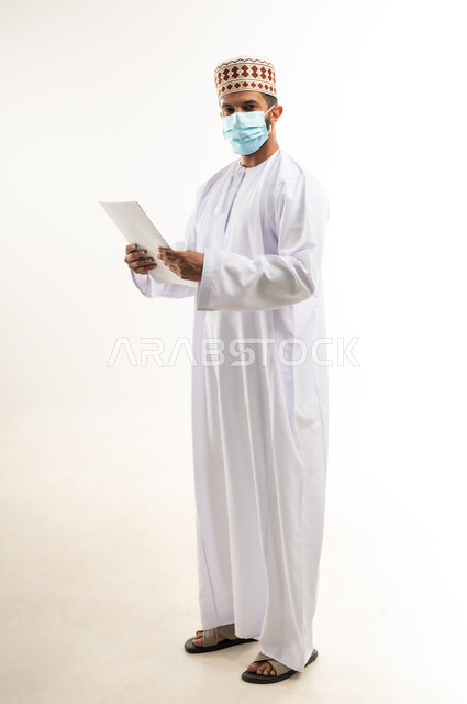 الحفاظ على السلامة من كوفيد 19، لوحات توعوية لتجنب إنتشار العدوى، اتباع الاجراءات الاحترازية للوقاية من فايروس كورونا، بورتريه لرجل عربي خليجي عماني يرتدي الدشداشة والكمة يضع الكمامة يمسك أوراق بيضاء فارغة, الجسم بالطول الكامل، خلفية بيضاء