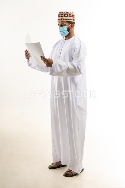 الحفاظ على السلامة من كوفيد 19، لوحات توعوية لتجنب إنتشار العدوى، اتباع الاجراءات الاحترازية للوقاية من فايروس كورونا، بورتريه لرجل عربي خليجي عماني يرتدي الدشداشة والكمة يضع الكمامة يمسك أوراق بيضاء فارغة, الجسم بالطول الكامل، خلفية بيضاء