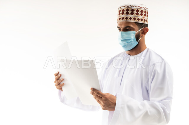 الحفاظ على السلامة من كوفيد 19، لوحات توعوية لتجنب إنتشار العدوى، اتباع الاجراءات الاحترازية للوقاية من فايروس كورونا، بورتريه لرجل عربي خليجي عماني يرتدي الدشداشة والكمة يضع الكمامة يمسك أوراق بيضاء فارغة, الجسم بالطول الكامل، خلفية بيضاء