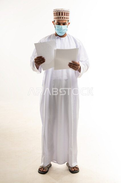 الحفاظ على السلامة من كوفيد 19، لوحات توعوية لتجنب إنتشار العدوى، اتباع الاجراءات الاحترازية للوقاية من فايروس كورونا، بورتريه لرجل عربي خليجي عماني يرتدي الدشداشة والكمة يضع الكمامة يمسك أوراق بيضاء فارغة, الجسم بالطول الكامل، خلفية بيضاء