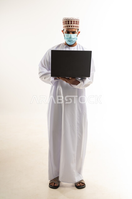 استخدام اللابتوب لانجاز المهام والعمل، بورتريه لرجل عربي خليجي عماني  يرتدي الدشداشة والكمة  يجلس على الكرسي يحمل الحاسوب المحمول، تطور التكنولوجيا والأجهزة التقنية، الجسم بالطول الكامل، خلفية بيضاء