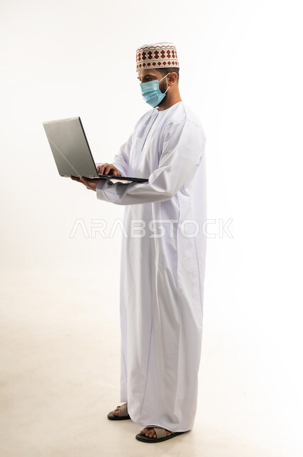 استخدام اللابتوب لانجاز المهام والعمل، بورتريه لرجل عربي خليجي عماني  يرتدي الدشداشة والكمة  يجلس على الكرسي يحمل الحاسوب المحمول، تطور التكنولوجيا والأجهزة التقنية، الجسم بالطول الكامل، خلفية بيضاء