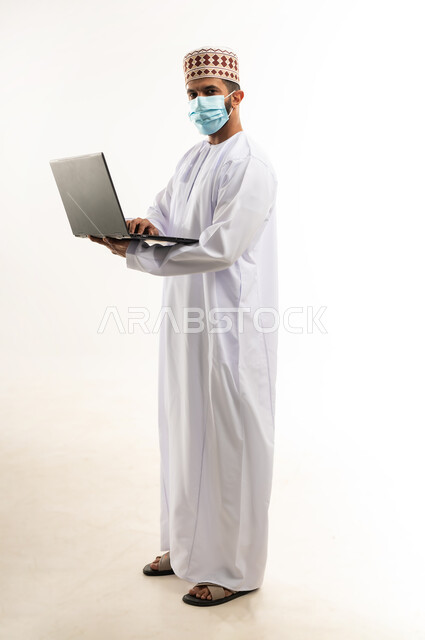 استخدام اللابتوب لانجاز المهام والعمل، بورتريه لرجل عربي خليجي عماني  يرتدي الدشداشة والكمة  يجلس على الكرسي يحمل الحاسوب المحمول، تطور التكنولوجيا والأجهزة التقنية، الجسم بالطول الكامل، خلفية بيضاء