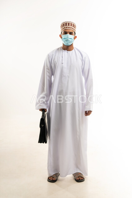 النظر الى الكاميرا بايماءات الجدية، الاهتمام بالاناقة والمظهر الخارجي، بورتريه لرجل أعمال عربي خليجي عماني يرتدي الدشداشة والكمة والكمامة الواقية يحمل حقيبة العمل، الالتزام باجراءات الوقاية من فايروس كورونا، صورة الجسم بالطول الكامل، خلفية بيضاء