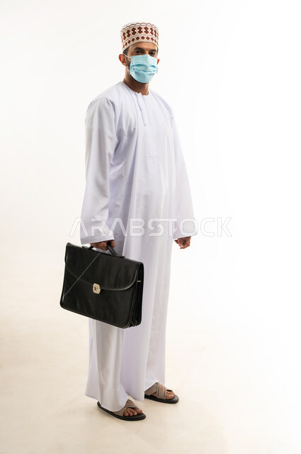 الاهتمام بالاناقة والمظهر الخارجي، بورتريه لرجل أعمال عربي خليجي عماني يرتدي الدشداشة والكمة والكمامة الواقية يحمل حقيبة العمل وينظر الى الكاميرا، الالتزام باجراءات الوقاية من فايروس كورونا، صورة الجسم بالطول الكامل، خلفية بيضاء