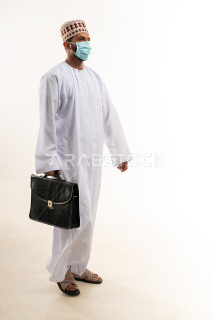 الاهتمام بالاناقة والمظهر الخارجي، بورتريه لرجل أعمال عربي خليجي عماني يرتدي الدشداشة والكمة والكمامة الواقية يحمل حقيبة العمل، الالتزام باجراءات الوقاية من فايروس كورونا، صورة الجسم بالطول الكامل، خلفية بيضاء