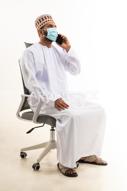 التواصل مع الاهل و الاصدقاء, بورتريه لشاب عربي خليجي عماني يرتدي الدشداشه و الكمه والكمامة يجلس على كرسي يقوم بإجراء مكالمة هاتفية عبر الهاتف المحمول، استخدام التقنيات الحديثة المتطورة, اتباع الاجراءات الوقائية من فايروس كورونا، خلفية بيضاء