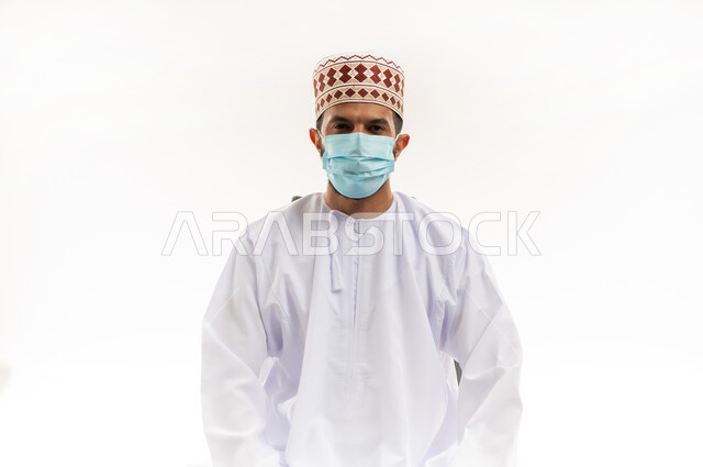 Looking at the camera with gestures of self-confidence, following precautionary measures to prevent the spread of infection, close-up portrait of a young Gulf Arab Omani man wearing a dishdasha and a kurta, wearing a medical mask to avoid infection, taking necessary measures to prevent the Corona virus, white background