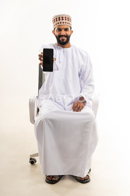 عرض شاشة سوداء فارغة على الجوال، استخدام الوسائل التقنية الحديثة، بورتريه لشاب عربي خليجي عماني يرتدي الدشداشة والكمه يحمل الهاتف المحمول ينظر الى الكاميرا بايماءات السرور، خلفية بيضاء