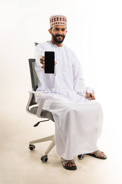 استخدام الوسائل التقنية الحديثة، عرض شاشة سوداء فارغة على الجوال،  بورتريه لشاب عربي خليجي عماني يرتدي الدشداشة والكمه يحمل الهاتف المحمول ينظر الى الكاميرا بايماءات السرور، خلفية بيضاء
