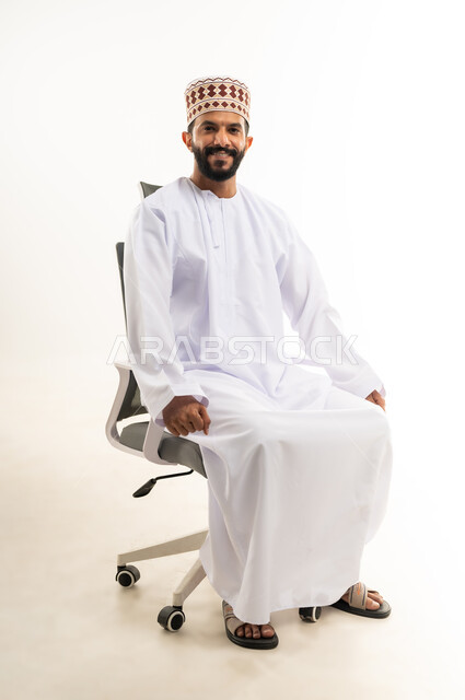 Concern for elegance and appearance, the concept of masculinity and self-confidence, taking personal photos for official documents, portrait of a smiling young Arab Gulf Omani man wearing a dishdasha and a keffiyeh sitting on a chair looking at the camera with gestures of pleasure and happiness, white background