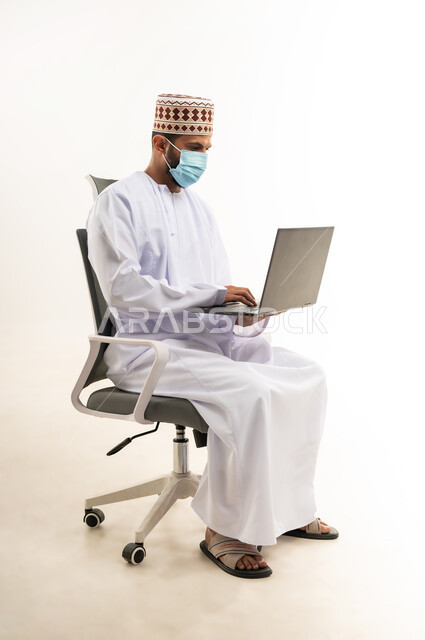 Using laptop to accomplish tasks and work, portrait of an Arab Gulf Omani man wearing a dishdasha and a turban sitting on a chair holding a laptop, development of technology and technical devices, white background