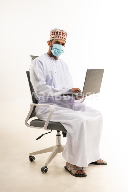 Using a laptop to accomplish tasks and work, portrait of an Arab Gulf Omani man wearing a dishdasha and a turban sitting on a chair holding a laptop and looking at the camera, development of technology and technical devices, white background