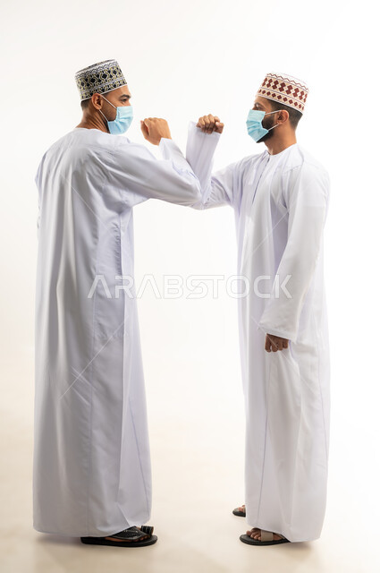 الحفاظ على المسافات وعدم التلامس، المصافحة الأمنة للإلتزام بتعليمات الصحة، بورتريه لموظفين عربيين خليجييين عمانييين يرتديان الدشداشة والكمة يتصافحان بالأكواع،  إرتداء القناع الطبي الوقائي للوقاية من فايروس كورونا كوفيد-19، الجسم بالطول الكامل، خلفية بيضاء