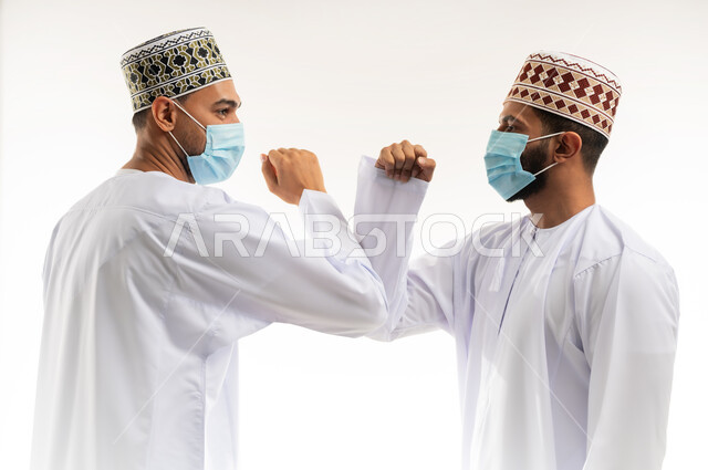 المصافحة الأمنة للإلتزام بتعليمات الصحة، بورتريه مقرب لشابين عربيين خليجييين عمانييين يرتديان الدشداشة والكمة يتصافحان بالأكواع،  إرتداء القناع الطبي الوقائي للوقاية من فايروس كورونا كوفيد-19، الحفاظ على المسافات وعدم التلامس، خلفية بيضاء