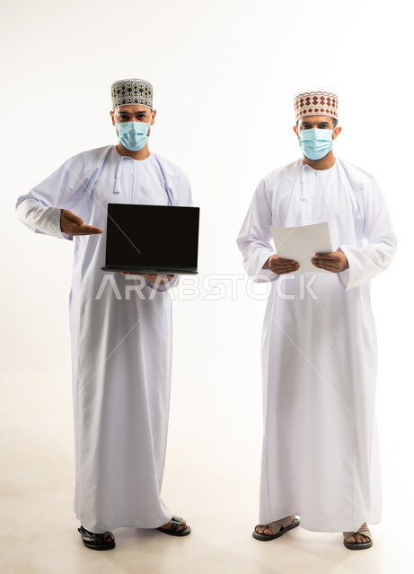 مفهوم الإدارة المتعاونة، بورتريه لموظفين عربيين خليجيين عمانيين يرتديان الدشداشة والكمة والكمامة يحملان الجهاز الحاسب والأوراق وينظران الى الكاميرا، اتباع الاجراءات الوقائية ضد كوفيد19, تحقيق أهداف الشركة وحل المشكلات، الجسم بالطول الكامل, خلفية بيضاء