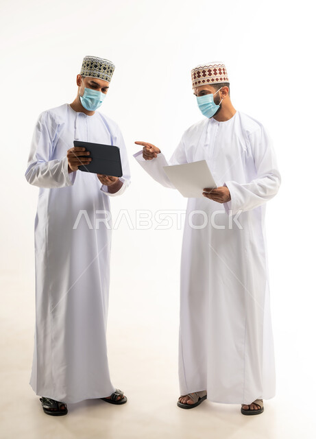 Achieving company goals and solving problems, collaborative management concept, portrait of two Gulf Arab Omani employees wearing dishdasha, kummah and mask discussing using tablet and papers, following preventive measures against Covid-19, full body, white background