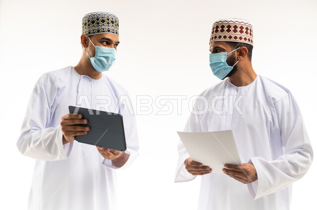 Collaborative management concept, close-up portrait of two Omani Gulf Arab employees wearing dishdasha, kummah and mask discussing using tablet and papers, following preventive measures against Covid19, achieving company goals and solving problems, full body, white background