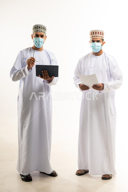 مفهوم الإدارة المتعاونة، بورتريه لموظفين عربيين خليجيين عمانيين يرتديان الدشداشة والكمة والكمامة يحملان جهاز التابليت والأوراق وينظران الى الكاميرا، اتباع الاجراءات الوقائية ضد كوفيد19, تحقيق أهداف الشركة وحل المشكلات، الجسم بالطول الكامل, خلفية بيضاء