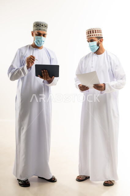 تحقيق أهداف الشركة وحل المشكلات، مفهوم الإدارة المتعاونة، بورتريه لموظفين عربيين خليجيين عمانيين يرتديان الدشداشة والكمة والكمامة يتناقشان باستخدام جهاز التابليت والأوراق، اتباع الاجراءات الوقائية ضد كوفيد19, الجسم بالطول الكامل, خلفية بيضاء