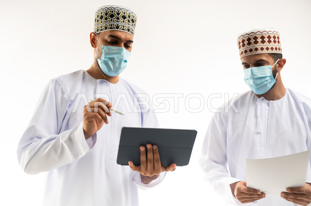 مفهوم الإدارة المتعاونة، بورتريه مقرب لموظفين عربيين خليجيين عمانيين يرتديان الدشداشة والكمة والكمامة يتناقشان باستخدام جهاز التابليت والأوراق، اتباع الاجراءات الوقائية ضد كوفيد19, تحقيق أهداف الشركة وحل المشكلات، الجسم بالطول الكامل, خلفية بيضاء