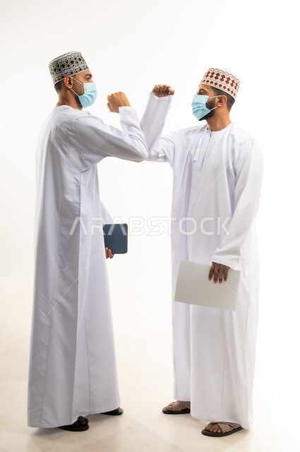 المصافحة الأمنة للإلتزام بتعليمات الصحة، بورتريه لموظفين عربيين خليجييين عمانييين يرتديان الدشداشة والكمة يتصافحان بالأكواع،  إرتداء القناع الطبي الوقائي للوقاية من فايروس كورونا كوفيد-19، الحفاظ على المسافات وعدم التلامس، الجسم بالطول الكامل، خلفية بيضاء