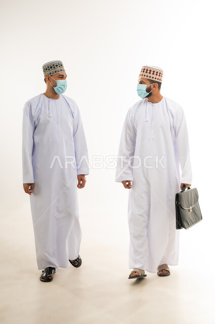 نقاشات وحوارات عمل بين رجال الاعمال، بورتريه لرجل عربي خليجي عماني يرتدي الدشداشة والكمة يحمل بيده حقيبة العمل ينظر الى زميله، ارتداء الكمامة الطبية واتباع الاجراءات الوقائية ضد فايروس كورونا، الجسم بالطول الكامل، خلفية بيضاء