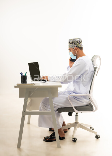 دمج التقنيات الحديثة بالحياة العملية, ارتداء الكمامة لاتخاذ الاجراءات الوقائية لفايروس كورونا, بورتريه من الجانب لرجل عربي خليجي عماني يرتدي الدشداشة والكمه يقوم باجراء مكالمة هاتفية عبر الهاتف المحمول، خلفية بيضاء