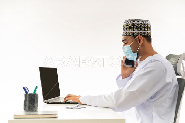 دمج التقنيات الحديثة بالحياة العملية, ارتداء الكمامة لاتخاذ الاجراءات الوقائية لفايروس كورونا, بورتريه مقرب من الجانب لرجل عربي خليجي عماني يرتدي الدشداشة والكمه يقوم باجراء مكالمة هاتفية عبر الهاتف المحمول، خلفية بيضاء